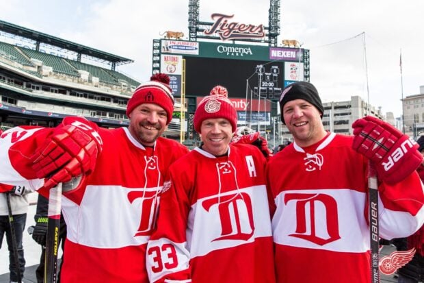 Three men wearing Detroit Red Wings jerseys and hats smiling at the camera