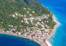 Coastal town with colorful houses, green hillside, rocky promontory, and turquoise ocean shoreline, scenic aerial view of a seaside community.