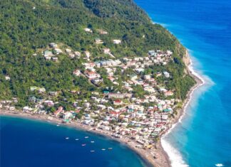 Coastal town with colorful houses, green hillside, rocky promontory, and turquoise ocean shoreline, scenic aerial view of a seaside community.