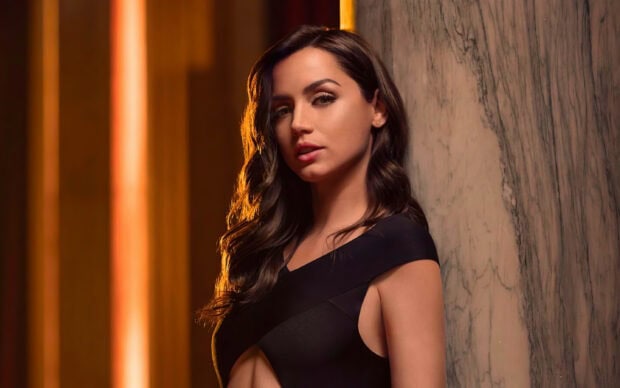 A woman poses confidently against a textured wall in a No Time To Die scene, 2K Desktop Wallpaper