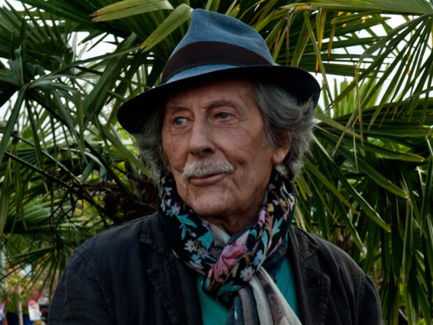 A thoughtful Jean Rochefort wearing a hat and floral scarf stands outdoors among green plants, HD Desktop Wallpaper