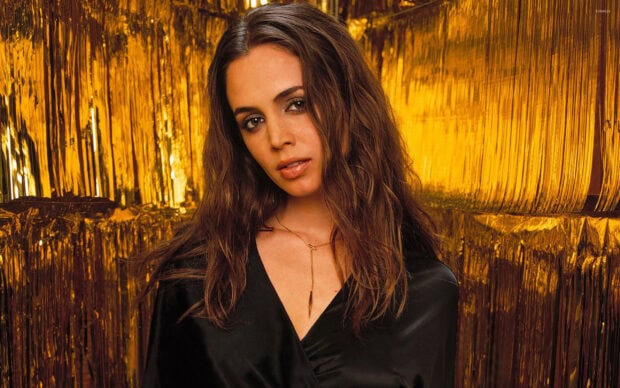 Eliza Dushku wearing a black outfit with long hair posing in front of a golden background