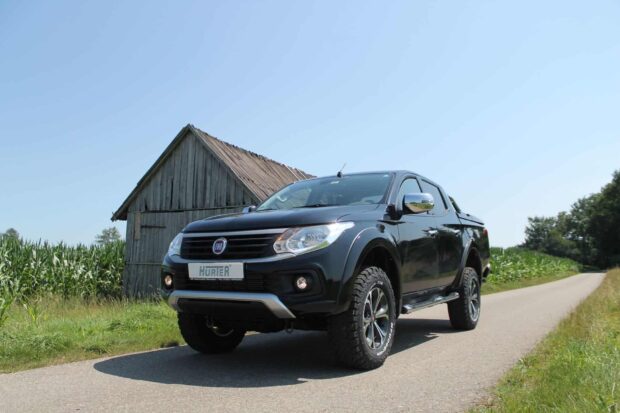 A black Fiat Fullback drives on a country road near a rustic wooden barn under clear skies, HD Desktop Wallpaper