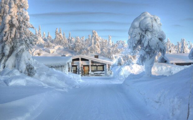 A snowy forest landscape featuring a cabin covered in thick snow surrounded by snow laden trees in Finland