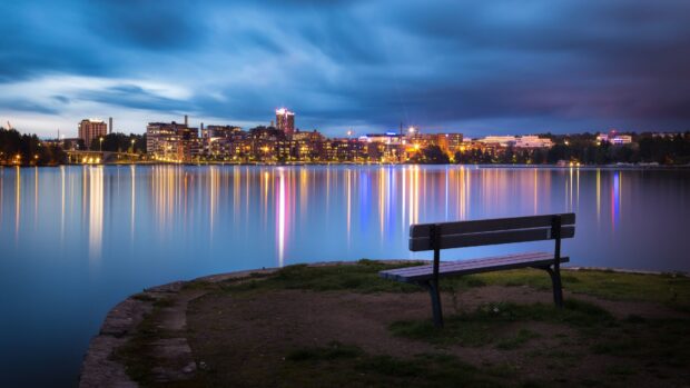 Calm waters reflecting city lights in Finland at dusk with a bench in the foreground