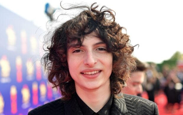Finn Wolfhard smiles warmly at an outdoor event with a blurred crowd in the background, 2K Desktop Wallpaper