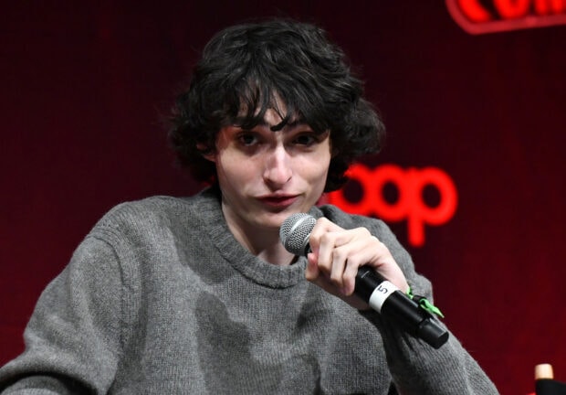 Finn Wolfhard is speaking into a microphone at an event with a red backdrop, HD Desktop Wallpaper