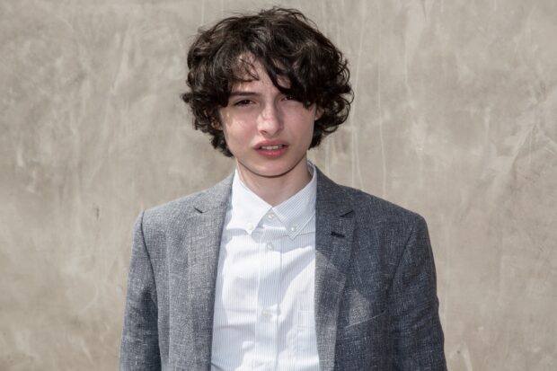 Finn Wolfhard is wearing a gray suit jacket and a striped white shirt standing against a light wall, 2K Desktop Wallpaper