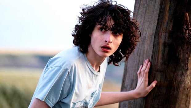 Finn Wolfhard is seen looking surprised while leaning on a wooden post in an outdoor setting, HD Desktop Wallpaper