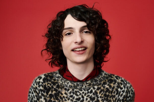 Finn Wolfhard is smiling wearing a patterned sweater against a red background, HD Desktop Wallpaper