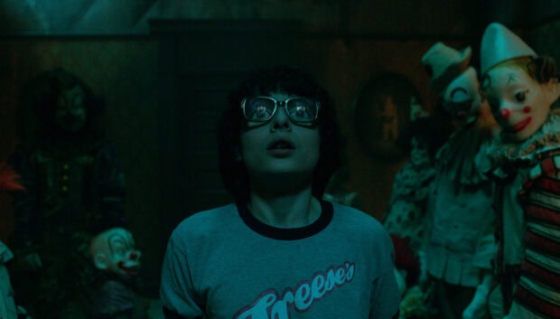 Finn Wolfhard looks surprised surrounded by creepy clown dolls in a dim room, HD Desktop Wallpaper