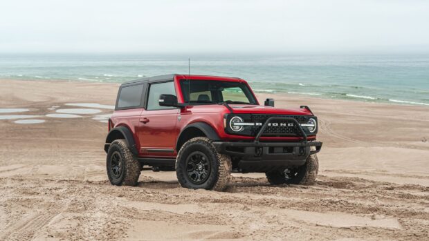 A red Ford Bronco parked on sandy beach near the ocean with clear sky in the background