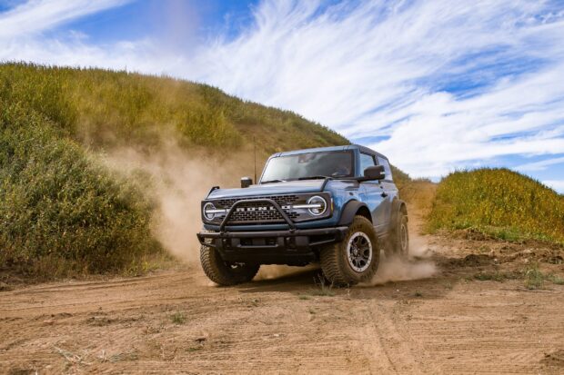 Blue Ford Bronco driving offroad on a dirt trail with grass and cloudy sky background