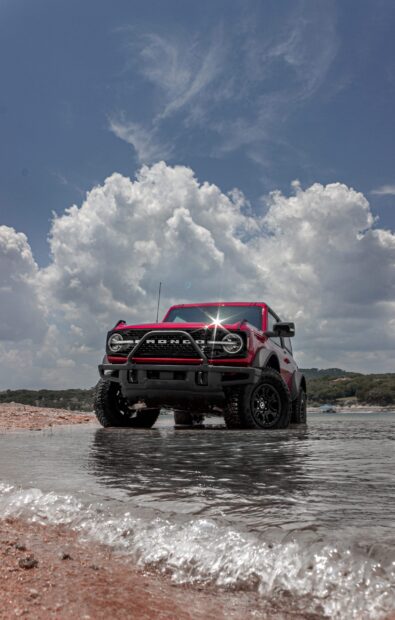 Red Ford Bronco driving through shallow water under cloudy sky