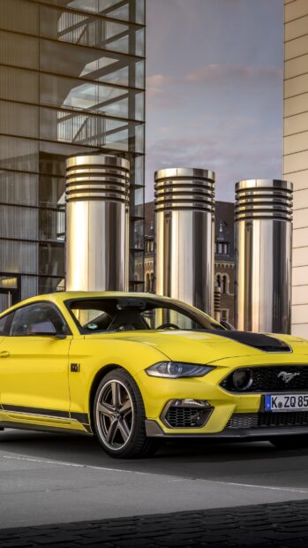 Bright yellow Ford Mustang parked in front of modern metallic structures at dusk