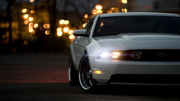 A white Ford Mustang car parked at night with headlights on and blurred city lights in the background