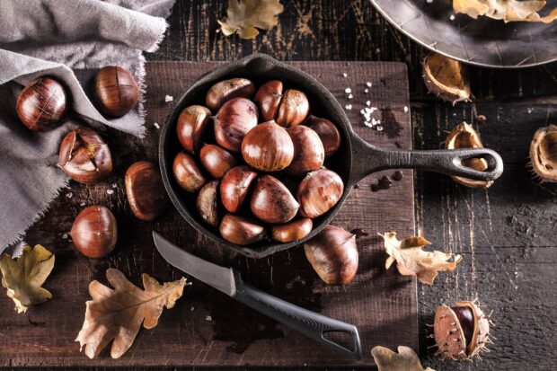 A cast iron pan filled with fresh roasted chestnuts surrounded by a knife and autumn leaves on a wooden cutting board, 2K Desktop Wallpaper