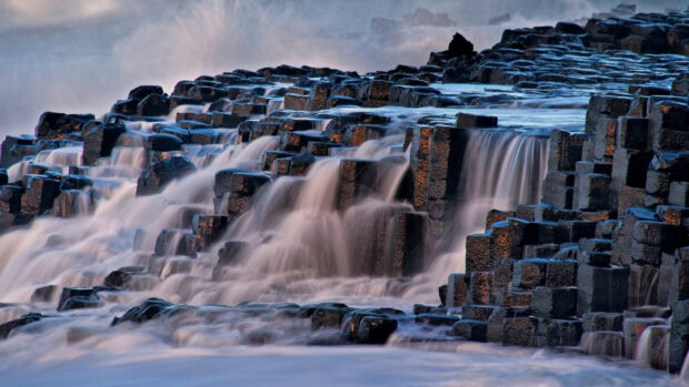 The image shows giant basalt columns with water cascading over them in Northern Ireland, 2K Desktop Wallpaper