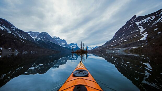 A kayak is floating on calm water surrounded by snowy mountains in Glacier National Park, 4K Desktop Wallpaper