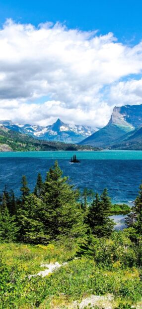 A scenic view of Glacier National Park showcases a turquoise lake surrounded by lush trees and snowy mountains, HD Phone Wallpaper