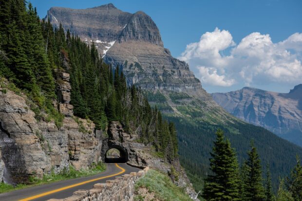 A winding mountain road passes through a rocky tunnel surrounded by green pine trees at Glacier National Park, HD Desktop Wallpaper