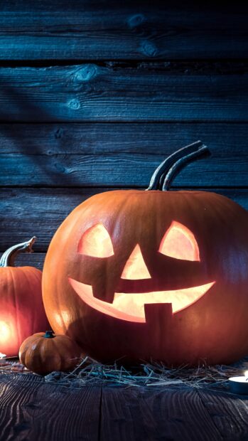 A glowing Halloween pumpkin with a carved smile sits on a wooden floor with other pumpkins, 4K Phone Wallpaper