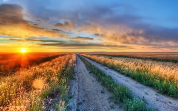 A golden sunset shines over a rural Montana landscape with a dirt road and fields, 2K Desktop Wallpaper