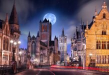The street showcases gothic architecture with illuminated historic buildings under a bright full moon, 4K Desktop Wallpaper