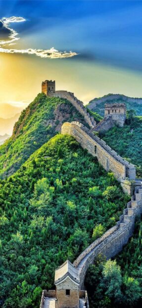 The Great Wall of China stretches over green hills under a clear sky with the sun setting