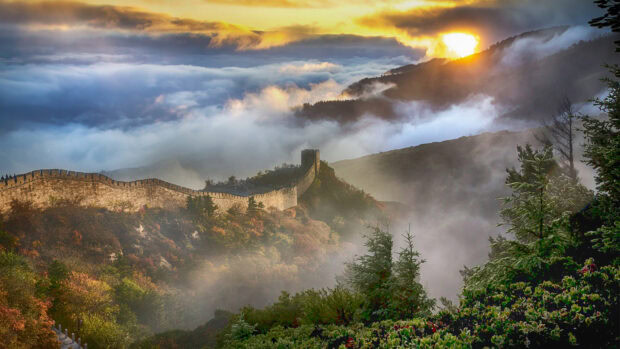 The Great Wall of China stretches across foggy mountains at sunrise with lush greenery surrounding it