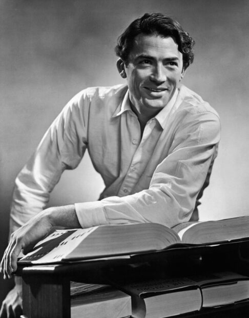 Gregory Peck is sitting by a table with open books smiling softly, HD Desktop Wallpaper