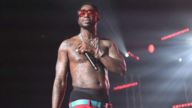 Gucci Mane performing shirtless with tattoos and jewelry on stage holding a microphone