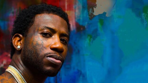 Close up of Gucci Mane with facial tattoos and jewelry against a colorful blue background