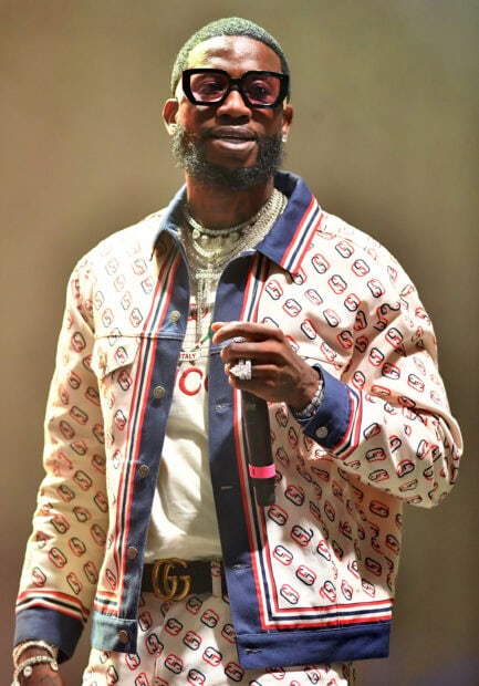Gucci Mane wearing a designer outfit and sunglasses holding a microphone on stage