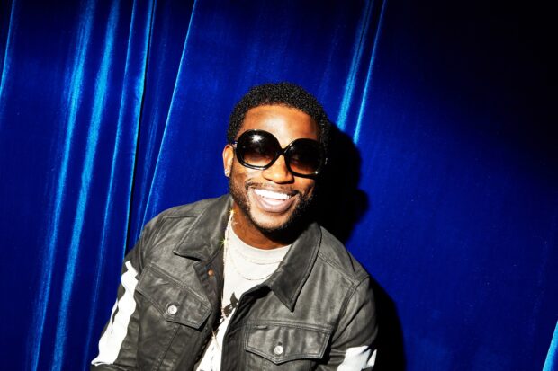 Gucci Mane wearing sunglasses and a jacket smiling against a blue backdrop