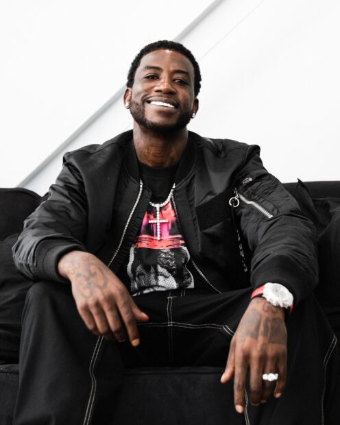 Gucci Mane smiling while sitting on a sofa showing his tattoos and jewelry