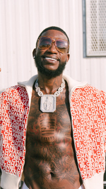 Gucci Mane smiling with sunglasses and a chain necklace against an outdoor wall