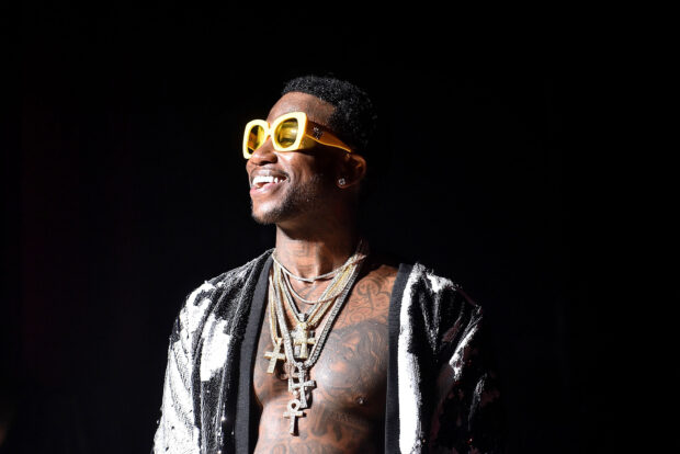 Gucci Mane wearing yellow sunglasses and multiple cross necklaces smiling on stage