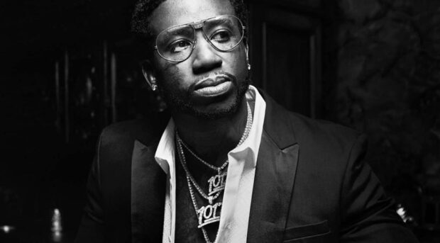 Gucci Mane wearing glasses and layered necklaces looking thoughtful in a black and white portrait