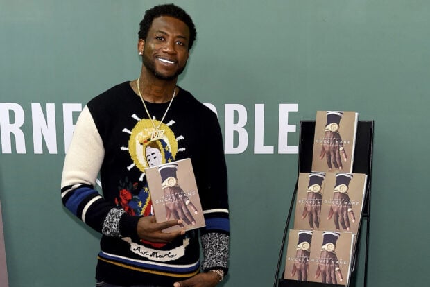 Gucci Mane holding a book with multiple copies of his autobiography displayed behind him