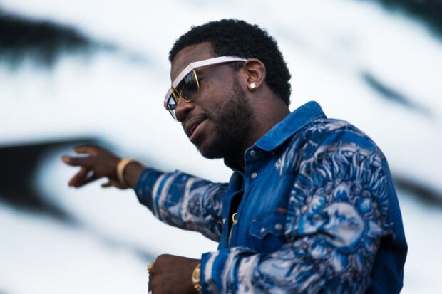 Gucci Mane wearing sunglasses and a patterned blue jacket with his arm extended on stage