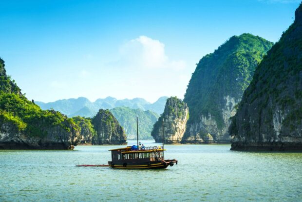 A traditional wooden boat sails through the limestone islands of Halong Bay in clear blue skies