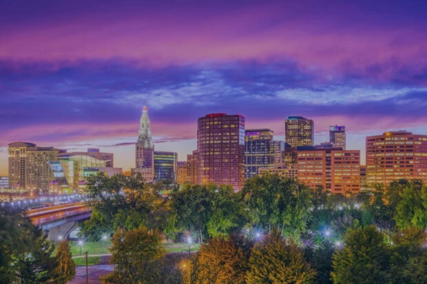 Hartford city skyline under a purple sunset sky with illuminated buildings and lush trees