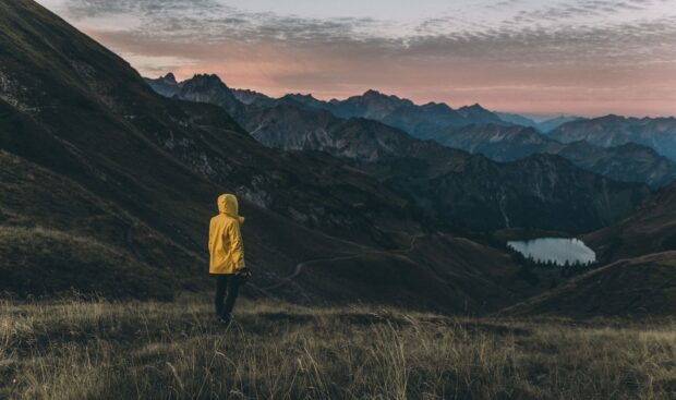 A hiker in a yellow jacket stands on a mountain trail surrounded by rugged peaks and a lake