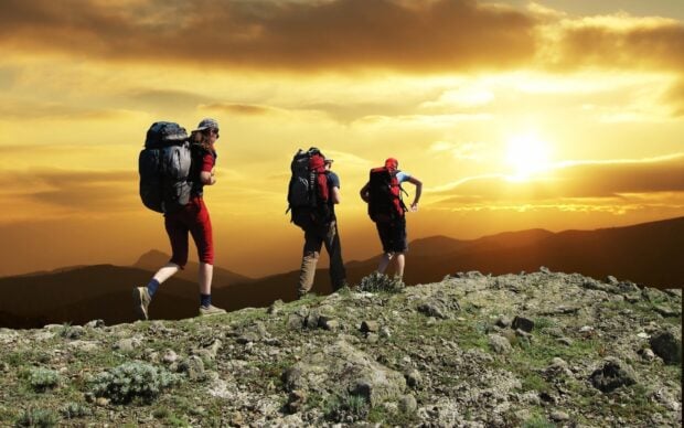 Three hikers with backpacks trekking on rocky terrain at sunset with mountains in the background