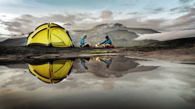 Two hikers sitting by a campfire next to a yellow tent reflecting on the water surface