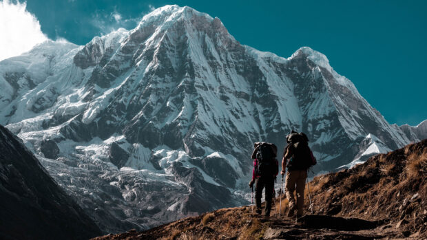 Two hikers trekking on a mountain trail with snowy peaks in the background