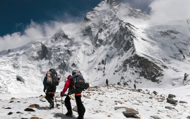 Two hikers trekking snowy mountain terrain with steep rocky peaks and cloudy sky