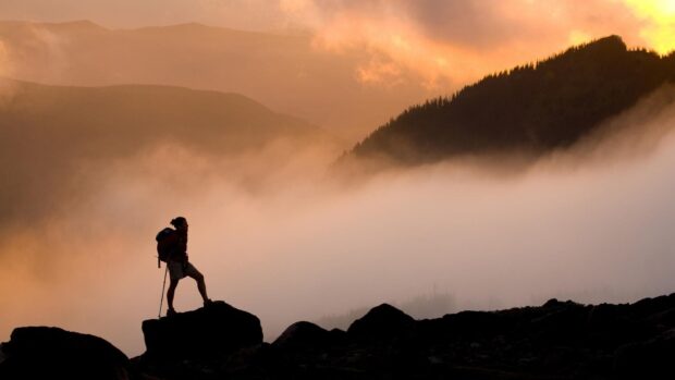 A hiker standing on rocks surrounded by fog and mountains at sunset