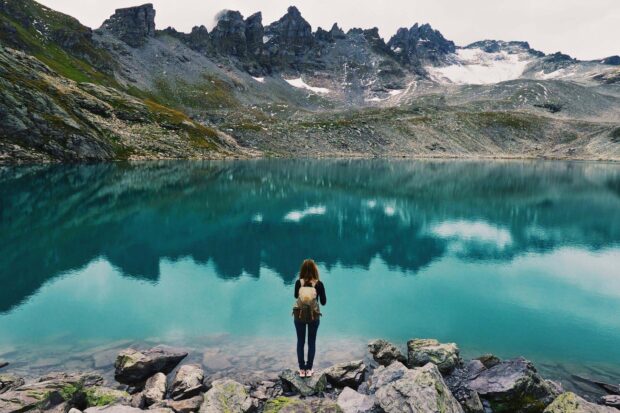 A hiker standing on rocky shore facing a mountain lake reflecting the alpine landscape